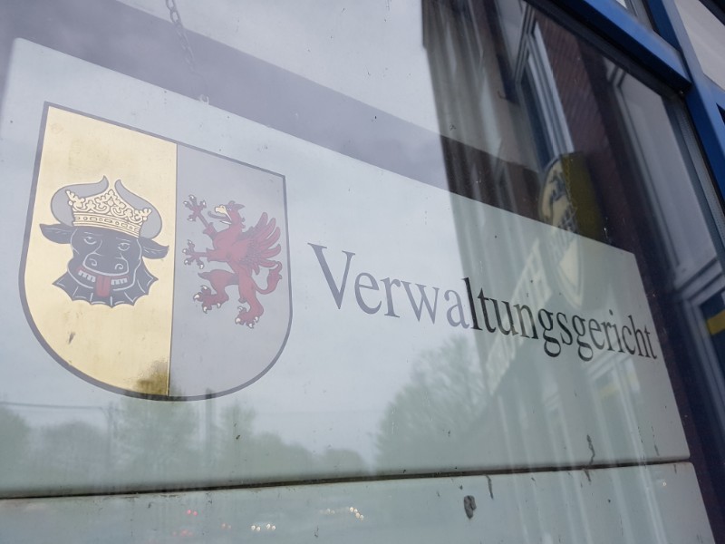 Das Foto zeigt hinter einer spiegelnden Glasscheibe ein Schild mit dem Aufdruck Verwaltungsgericht, rechts davon das Wappen des Bundeslandes Mecklenburg-Vorpommern.