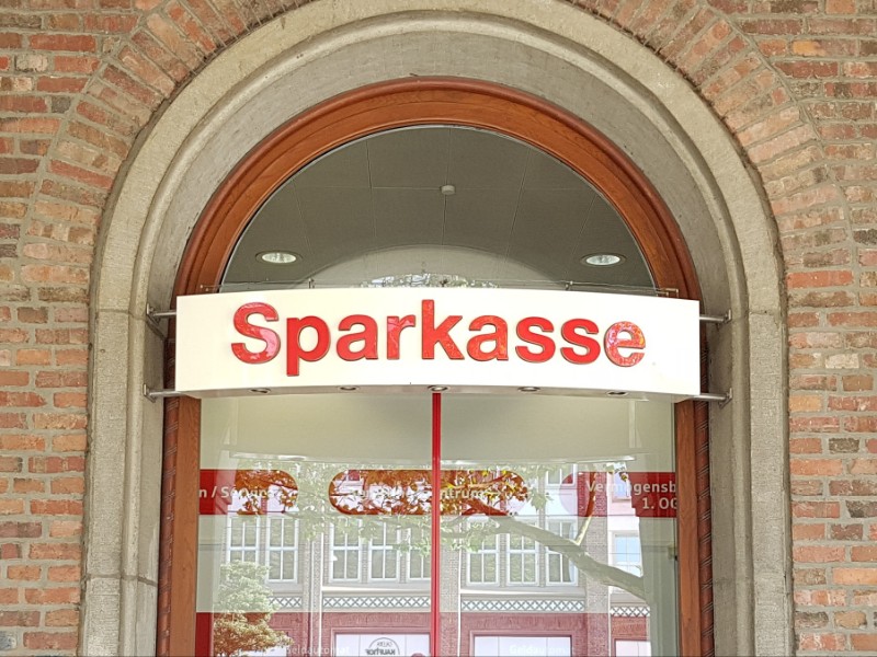 Das Foto zeigt den Eingangsbereich einer Sparkasse, über der Tür befindet sich ein Schild mit der Aufschrift Sparkasse, rot auf weißem Grund.