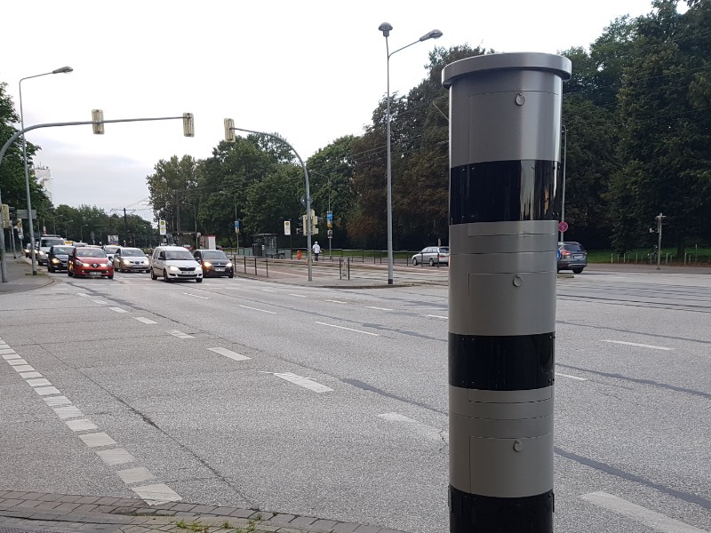 Das Foto zeigt ein stationäres Blitzgerät, im Hintergrund Straßenverkehr.