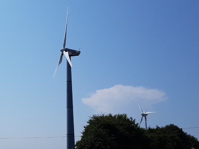 Das Foto zeigt zwei Windkraftanlagen, blauen Himmel und ein Stück Wald.