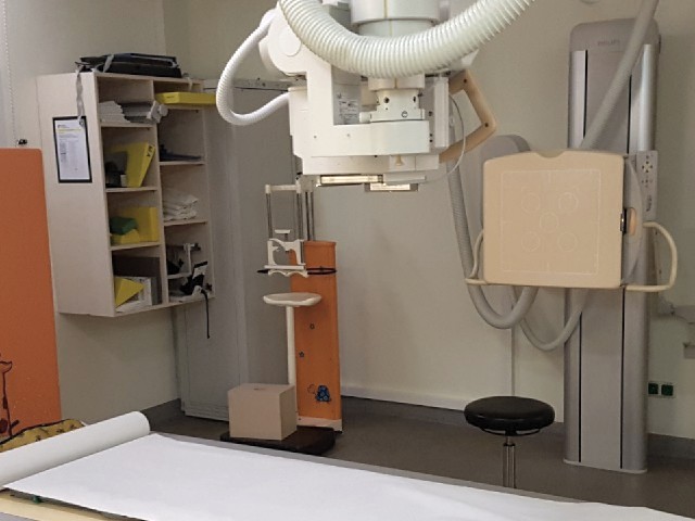 Auf dem Foto ist der Blick in ein Behandlungszimmer mit Röntgengerät und Liege zu sehen, im Hintergrund befindet sich ein Regal mit Zubehör.